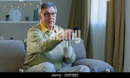 Grauhaariger alter Mann mit Brille, der auf der Couch sitzt und sich zu Hause entspannt, Tee trinkt, Kaffee trinkt, alter Mann verwendet die Fernbedienung, um umzuschalten Stockfoto