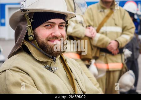 Porträt eines bärtigen Retters am Sommertag auf der Straße. Feuerwehrmann in Schutzkleidung und Helm schaut in die Kamera und lächelt. Authentischer weißer Rettungshelfer. Positive Emotionen... Stockfoto