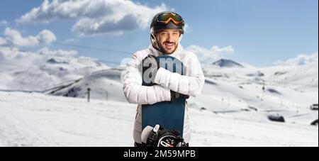 Junger Mann, der ein Snowboard umarmt und auf einem verschneiten Berg posiert Stockfoto