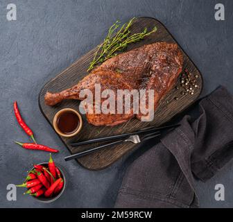 Gegrillte Ente oder Hähnchen in Gewürzen und Marinade auf einem Holzbrett mit frischen Gewürzen und Chili-Pfeffer auf dunklem Hintergrund. Draufsicht und Kopierbereich. Stockfoto