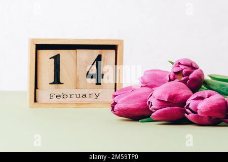Ein Haufen lila Tulpen und ein Holzblock-Kalender mit Datum vom 14. februar auf einem Tisch. Valentinstag-Urlaubskonzept. Nahaufnahme Stockfoto