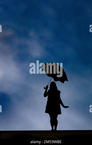 Ein Mädchen steht mit Sonnenschirm in der rechten Hand und blickt auf die Kamera, die Silhouette des Himmels Stockfoto