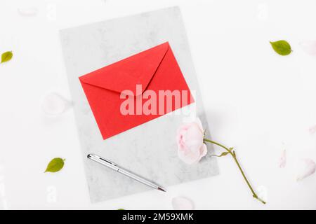 Roter Papierumschlag mit leerer Notiz und Valentinsrosa auf weißem Hintergrund. Flach liegend, Draufsicht romantischer Liebesbrief Stockfoto