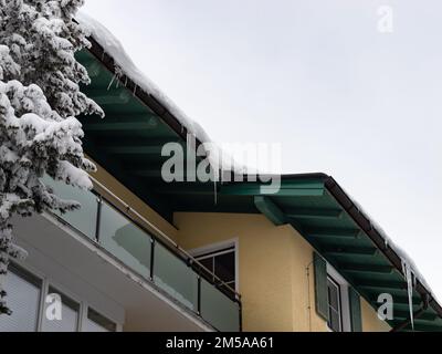 Schnee und Eiszapfen auf einem Holzdach. Mit Blick auf die Dachrinne eines gelben Gebäudes mit Balkon. Der schwere Schnee liegt auf dem Dach. Stockfoto