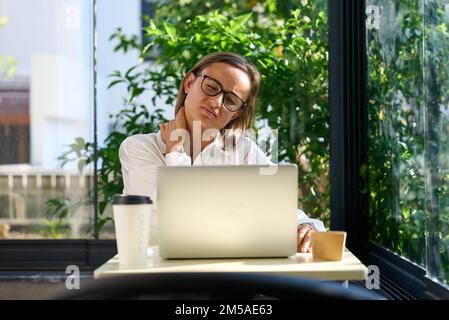 Müde gestresste Frau mit Nackenschmerzen, die im Home Office am Tisch sitzt. Überarbeitete junge Dame massiert Nackenschmerzen Stockfoto