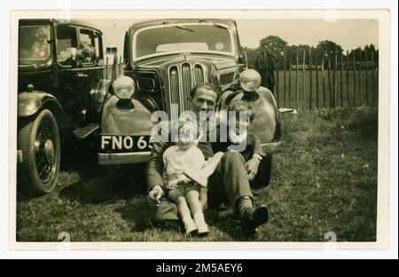 Originale, charmante Postkarte aus der Zeit der späten 1930er Jahre, auf der zwei kleine Kinder vor einem Ford Anglia sitzen, an einem Tag mit ihrem Vater, vielleicht einer Rallye. Canterbury und Umgebung, Kent, England, Großbritannien. Stockfoto