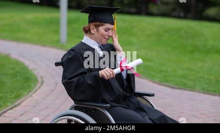 Eine weiße Frau im Rollstuhl hält ihr Diplom und weint vor Freude draußen. Stockfoto