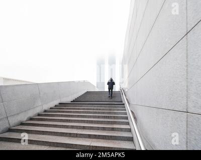 Der Mann geht nach oben über eine Fußgängerüberquerung. Die Wolkenkratzer sind im Nebel. Einsamkeit in der Großstadt. Urbane Architektur und einsamer Mann. Stockfoto