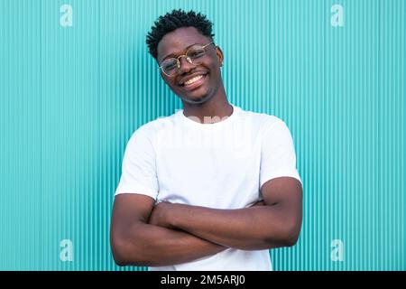Fröhlicher junger afroamerikanischer Mann, der die Arme überkreuzte, auf türkisfarbenem Hintergrund - Porträt eines fröhlichen jungen Mannes - Happines und positives Konzept Stockfoto