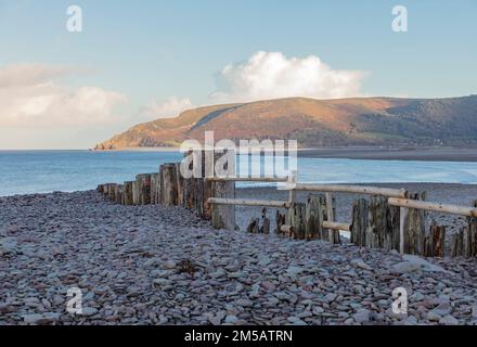 Mit Blick über einen Kieselstrand mit alten Holzzäunen und in der Ferne Sonnenschein auf Küstenhügeln Stockfoto
