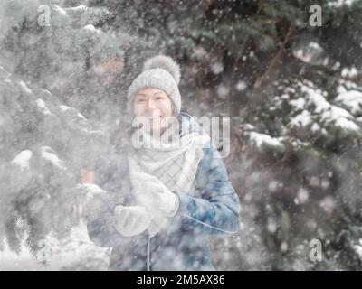 Eine lächelnde Frau in einem gestrickten Hut spielt mit Schnee. Spaß im Park zwischen schneebedeckten Tannen. Die Frau lacht, während sie Schneeball wirft. Kalte Jahreszeit. Stockfoto