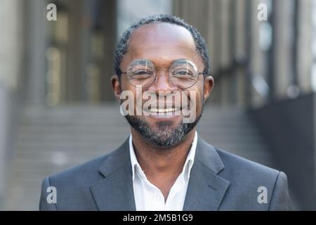 Nahaufnahme Foto eines erfolgreichen und lächelnden afroamerikanischen Mannes, eines Geschäftsmanns mit Brille, der lächelt und von außerhalb des Bürogebäudes auf die Kamera schaut. Stockfoto