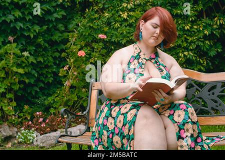Weiße argentinische Latina-Frau in Übergröße, die ein Buch liest, das auf einer Holzbank im Garten sitzt, mit einer grünen Wand im Hintergrund und Kopie Stockfoto
