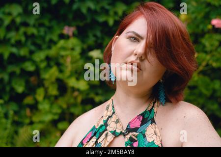 Porträt einer wunderschönen jungen Latina-Kaukasierin in Übergröße, kurze rote Haare, gekleidet in einem Blumenkleid, entspannt und ruhig in einem Garten Stockfoto