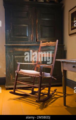 Antiker Schaukelstuhl aus Holz neben einem grün bemalten Schrank im Speisesaal mit gelb bemalten Dielen in einem alten Haus im französischen Stil von 1722. Stockfoto