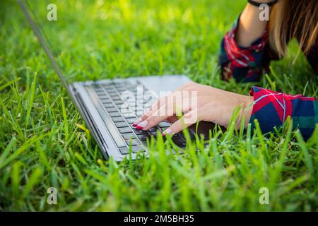 Nahaufnahme der Hände eines Mädchens, die mit einem Laptop tippen, der auf einer Wiese ruht, Technologie im Freien Stockfoto