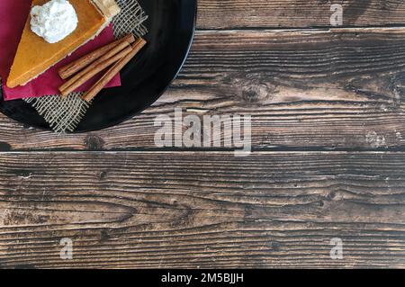 Ein Stück Kürbiskuchen auf einem schwarzen runden Teller in der oberen linken Ecke eines hölzernen Hintergrunds Stockfoto