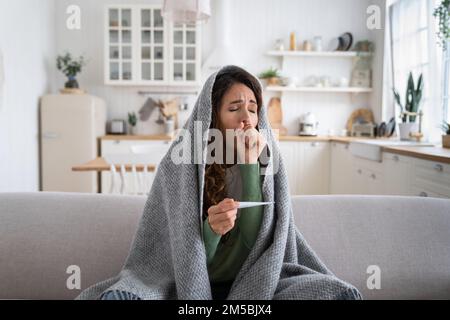 Eine kranke Frau hustet, misst die Temperatur mit einem Thermometer, sitzt auf dem Sofa und ist in eine Decke gewickelt Stockfoto