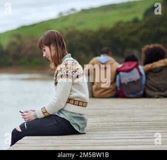 Ich genieße einen Moment für mich. Eine junge Frau, die eine SMS schickt, während sie am Pier am See sitzt. Stockfoto