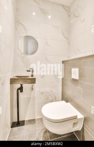 Eine weiße Toilette in einem kleinen Badezimmer mit Marmorfliesen an den Wänden und einem runden Spiegel darüber Stockfoto