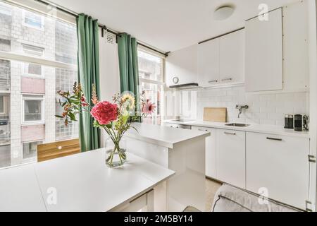 Eine weiße Küche mit Blumen in der Vase auf der Theke und Fenster auf der linken Seite, mit Blick auf die Straße Stockfoto