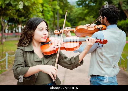 Zwei junge Violinisten, die in einem Park Geige spielen. Porträt von Mann und Frau, die im Park Geige spielen. Violinisten-Mann und -Frau zurück Stockfoto