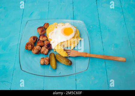 Rumänische traditionelle Speisen, gebratene Würstchen, weiche Eier und „Mamaliga“, Nahaufnahme von der Seite, auf blauem Hintergrund Stockfoto