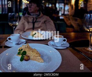 Napoleonkuchen auf einem weißen Keramikteller und Kaffeetassen, Gläser mit unvollendetem Weißwein und eine ziemlich reife Frau, die im Bac aus dem Fenster schaut Stockfoto