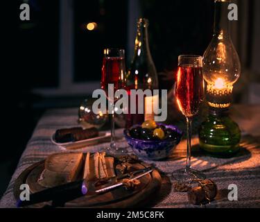 Zwei Gläser mit Wein, Oliven in einer blauen Keramikschüssel, Käse mit Nüssen auf dem Tisch in den Strahlen einer Kerosinlampe. Stockfoto