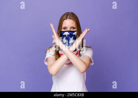 Junge wunderschöne patriotische Frau trägt die US-Flagge Gesichtsmaske auf violettem Hintergrund mit gekreuzten Armen und macht ein Stoppschild mit einem ernsthaften und selbstbewussten Ausdruck, Schutzgeste Stockfoto