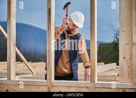 Ein Mann, der ein Holzrahmenhaus baut. Zimmermann hämmert Nagel in Holzbrett mit Hammer. Zimmereikonzept. Stockfoto