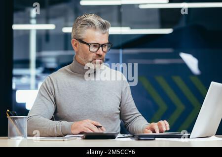 Ein seriöser Geschäftsmann im Büro, der mit einem Laptop arbeitet, ein grauhaariger Senior-Mann, der am Schreibtisch sitzt. Stockfoto