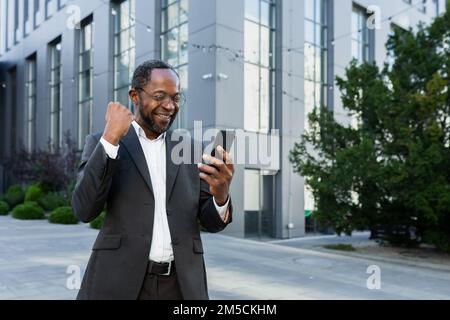 afroamerikanischer Geschäftsmann Chef vor dem modernen Bürogebäude mit Telefon, Senior Mann feiert Siegeserfolg, liest gute Nachrichten online vom Smartphone, hält Hand-hoch-Geste des Triumphes. Stockfoto
