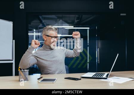 Erfolgreicher, grauhaariger Boss, der in einem modernen Büro arbeitet, ein erwachsener Geschäftsmann, der den Sieg feiert, auf dem Bildschirm steht und zufrieden mit seiner Leistung ist, ein leitender Mann, der lächelt und tanzt. Stockfoto