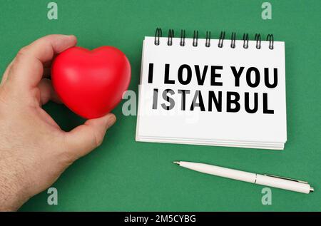 Auf der grünen Oberfläche ist ein Stift, ein Notizblock mit der Inschrift - Ich liebe dich Istanbul. Ein Mann hat ein Herz in der Hand. Stockfoto