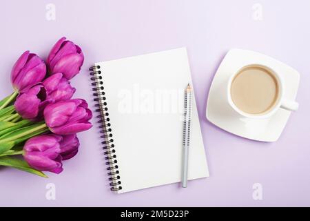 Leeres Notizbuch, Stift, Kaffeetasse und lila Tulpen auf lila Hintergrund. Draufsicht, flaches Lay, Modell Stockfoto