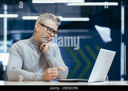 Der grauhaarige Geschäftsmann im Büro war frustriert und traurig, als er im Online-Shop Banktransaktionen und Einkäufe tätigte, ein Mann, der eine Kreditkarte hatte und ein Notebook benutzte. Stockfoto