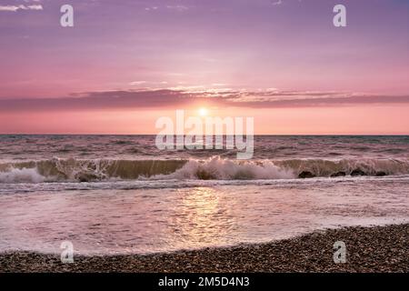 Das Meer sprudelnde Wellen beleuchtet von der untergehenden Sonne vor dem wunderschönen Sonnenuntergang am Himmel. Getöntes Bild in trendy magentafarbener Farbe des Jahres 2023. Naturhintergrund. Großartiges Tapetendesign. Speicherplatz kopieren. Stockfoto