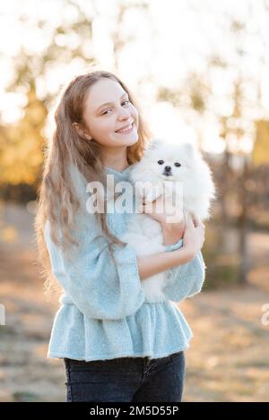 Süßes lächelndes Teenager-Mädchen 15-16 Jahre alt, das den weißen, flauschigen spitz Welpen-Hund über den gelben Herbsthintergrund im Park hält. Freundschaft. Stockfoto