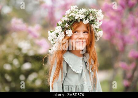 Niedliches, lachendes kleines Mädchen im Alter von 3-4 Jahren mit langen lockigen roten Haaren, Blumenkranz und stilvollem rustikalem Kleid über dem Naturhintergrund im Garten Outdo Stockfoto