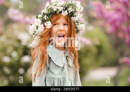 Niedliches, lachendes kleines Mädchen im Alter von 3-4 Jahren mit langen lockigen roten Haaren, Blumenkranz und stilvollem rustikalem Kleid über dem Naturhintergrund im Garten Outdo Stockfoto