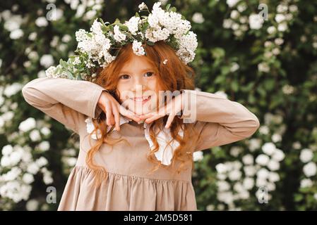 Niedliches, lachendes kleines Mädchen im Alter von 3-4 Jahren mit langen lockigen roten Haaren, Blumenkranz und stilvollem rustikalem Kleid über dem Naturhintergrund im Garten Outdo Stockfoto