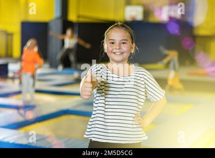 Fröhliches Tween-Mädchen, das im aufblasbaren Vergnügungspark mit den Daumen nach oben auf Trampoline zeigt Stockfoto