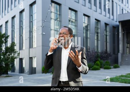 Wütender afroamerikanischer Boss streitet und schreit, während er telefoniert, Senior-Mann im Business-Anzug außerhalb des Bürogebäudes. Stockfoto