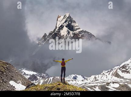 Wandern Sie in einem gelben Hemd mit Rucksack, der mit erhobenen Händen auf dem Felsen steht und den Blick auf die verschneiten Berge an sonnigen Tagen genießt Stockfoto