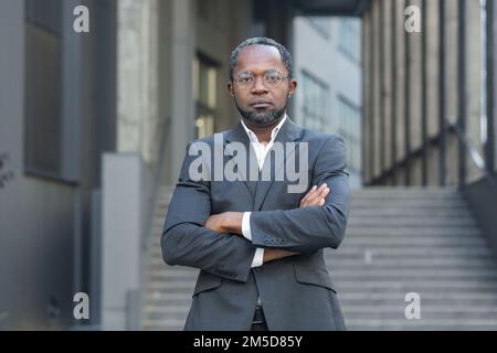 Seriöser und selbstbewusster afroamerikanischer Mann im Business-Anzug, der vor die Kamera schaut, Geschäftsmann mit gekreuzten Armen außerhalb des Bürogebäudes. Stockfoto