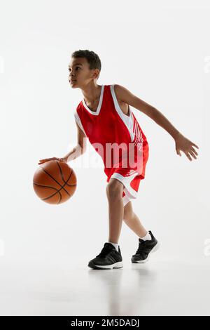 Porträt eines Jungen in roter Uniform, der Basketball mit grauem Studiohintergrund spielt. Dribbling-Übungen Stockfoto