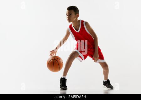 Porträt eines Jungen in roter Uniform, der Basketball spielt, über grauen Studiohintergrund dribbelt. Motivierter Sportler Stockfoto