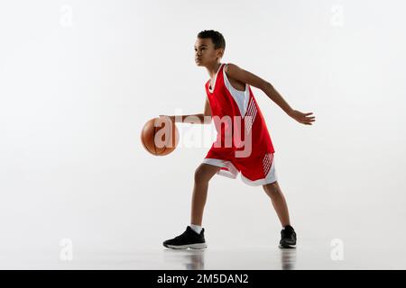 Porträt eines Jungen in roter Uniform, der Basketball spielt, über grauen Studiohintergrund dribbelt. Konzentration Stockfoto