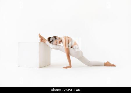 Junge Frau, die Yoga praktiziert, eine Variation von Hanumanasana-Training durchführt, eine Längsspaltung, wobei ihr Bein auf einem Würfel ruht, trainiert weißes spo Stockfoto